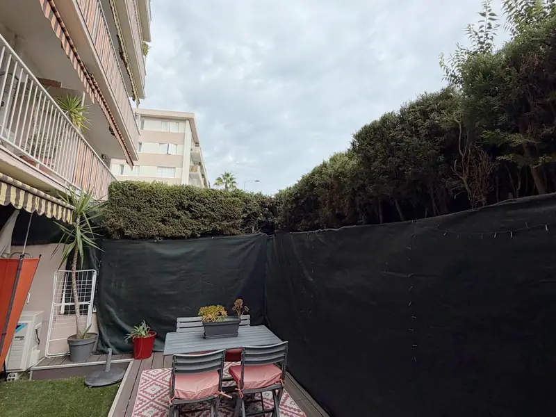 Terrasse en bois d'un rez-de-jardin : table, chaises et tapis, haie de conifères taillée au-dessus d'un brise-vue noir