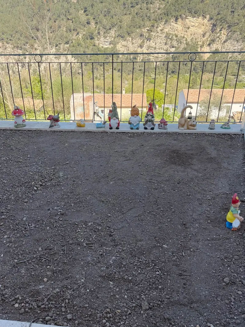 Avant : terrasse de terre nue nivelée, bordée d'un garde-corps en fer forgé, falaise boisée au fond